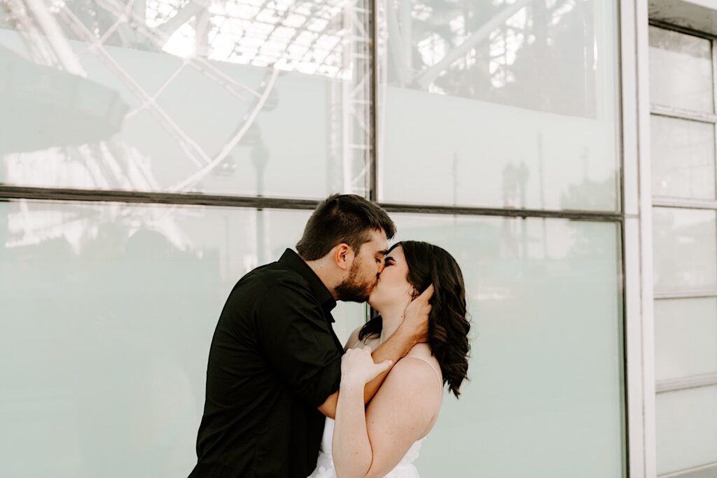 During their engagement session a couple kiss and embrace while standing in front of a window with the reflection of the Ferris Wheel at Navy Pier in Chicago on it