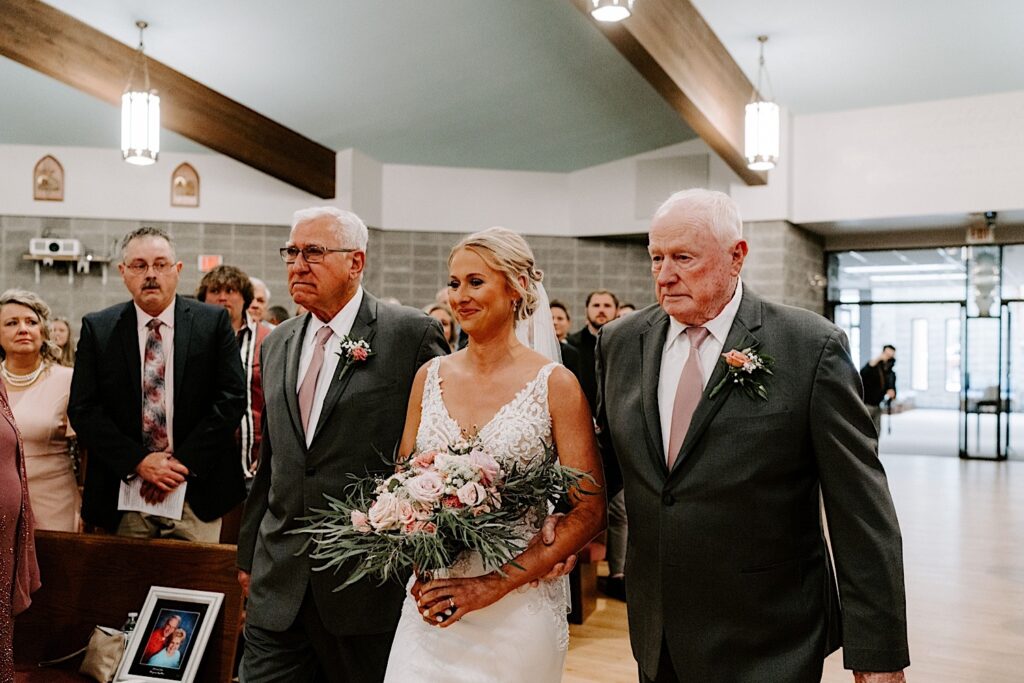 A bride smiles as she is escorted down the aisle of her wedding by her two grandfathers