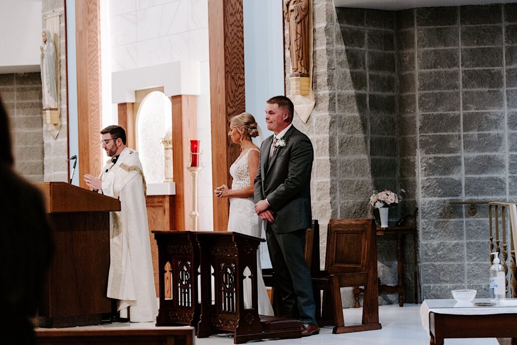 A bride and groom stand while a priest gives a sermon during their wedding ceremony