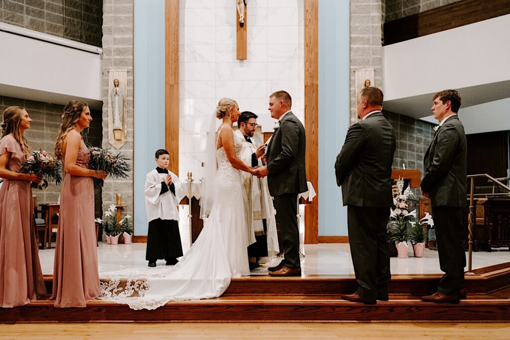 A bride and groom hold hands while their wedding parties stand on either side of them during their wedding ceremony, the pastor is speaking while standing in between them
