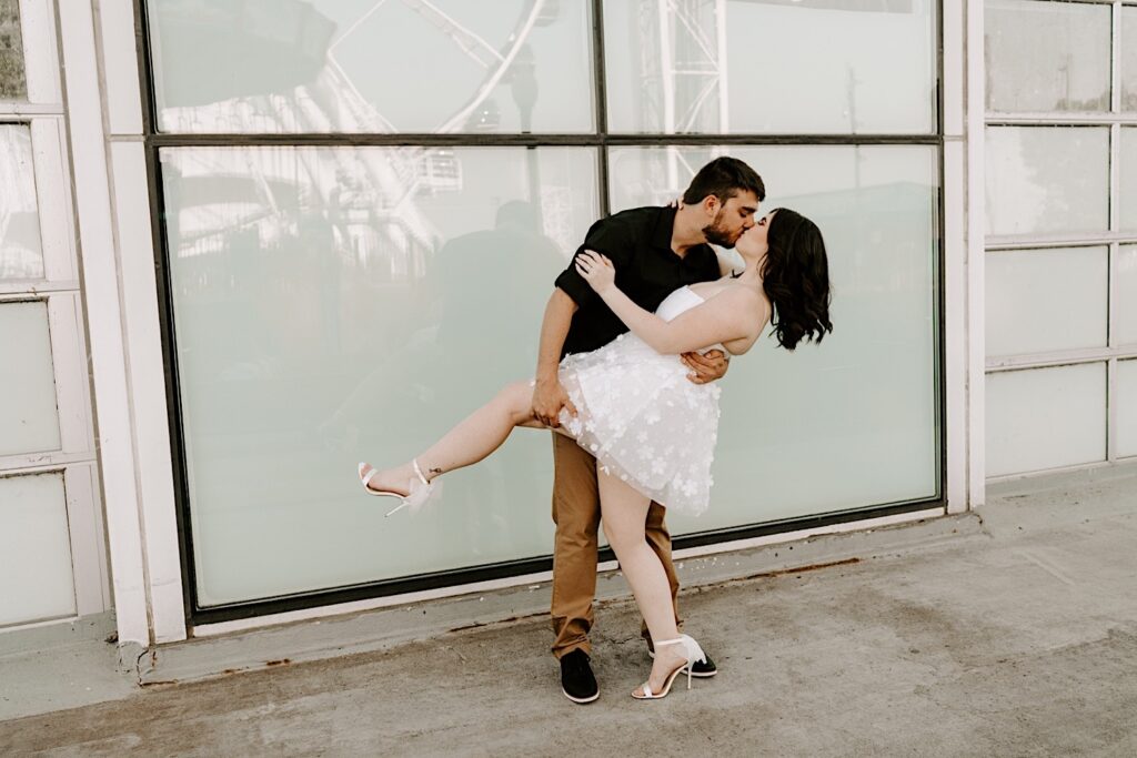 A man kisses and dips his fiancée during their engagement session at Navy Pier in Chicago, behind them is a window with the Ferris Wheel's reflection in it