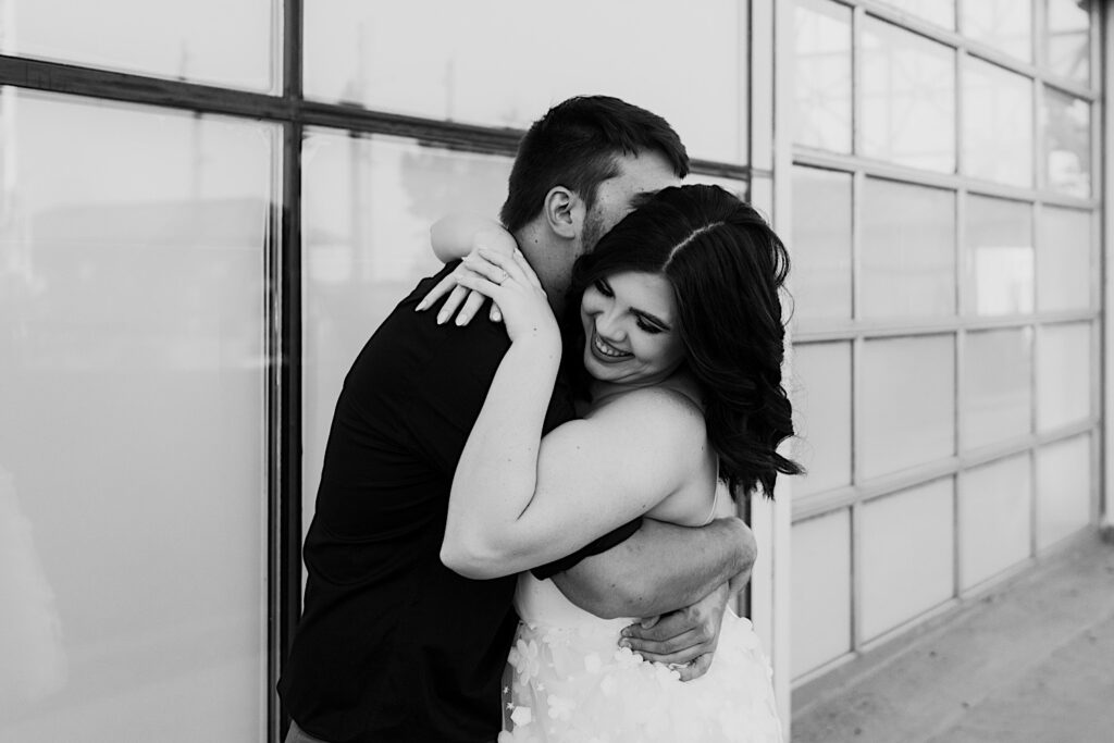 Black and white photo of a woman smiling as she is hugged by her fiancé in front of a window