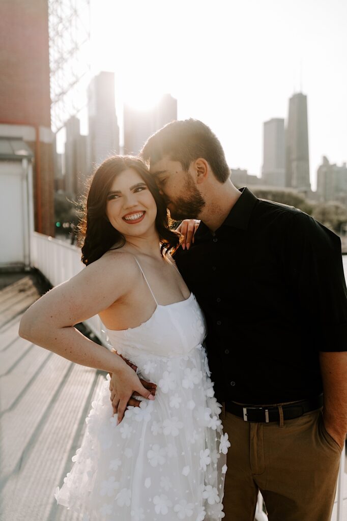 A woman smiles at something off camera as her fiancé whispers something in her ear with the Chicago skyline in the background