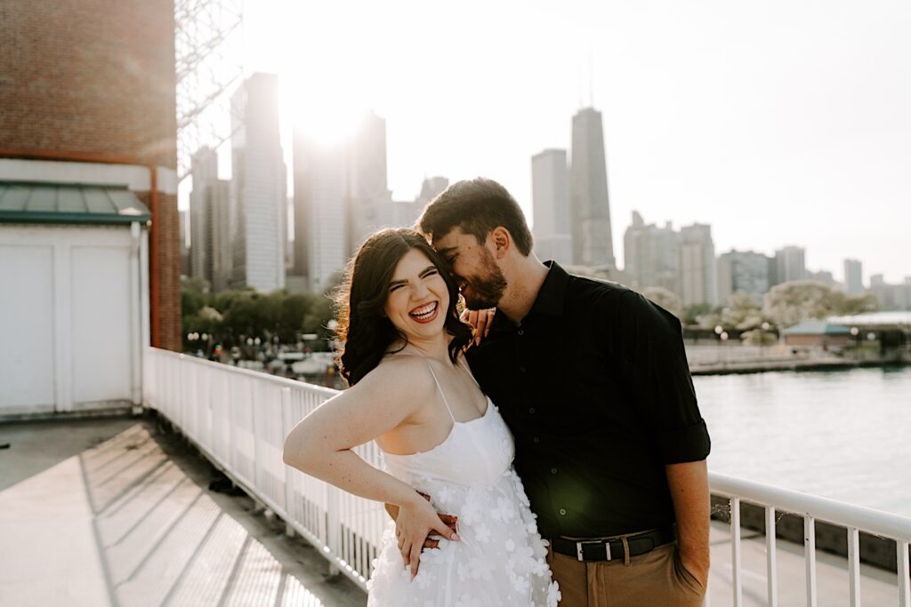 During their engagement session at Navy Pier a woman smiles at the camera as her fiancé whispers something in her ear with the Chicago skyline in the background