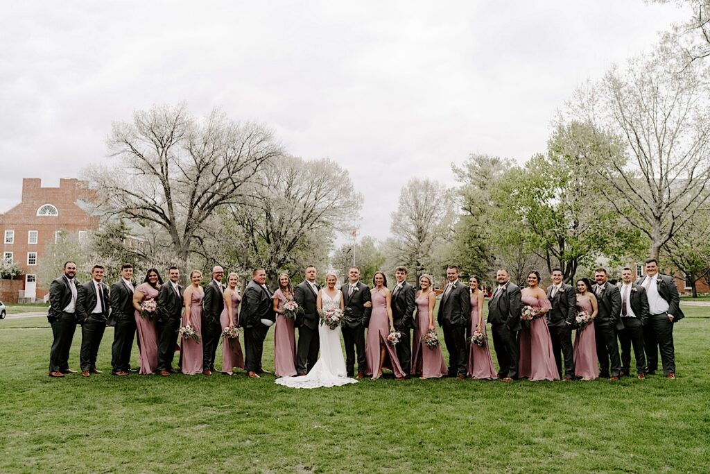 A bride and groom stand in a field with their wedding parties on either side of them, everyone is smiling and looking at the camera