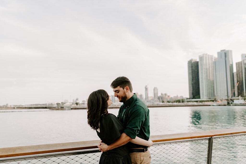 A couple embrace and look at one another while standing next to a railing at Navy Pier looking out over the Chicago skyline and Lake Michigan