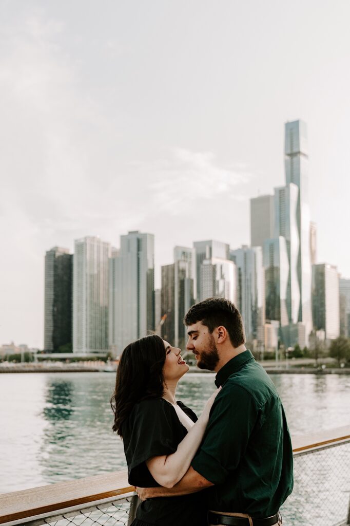 A couple embrace and look at one another while standing next to a railing at Navy Pier looking out over the Chicago skyline and Lake Michigan