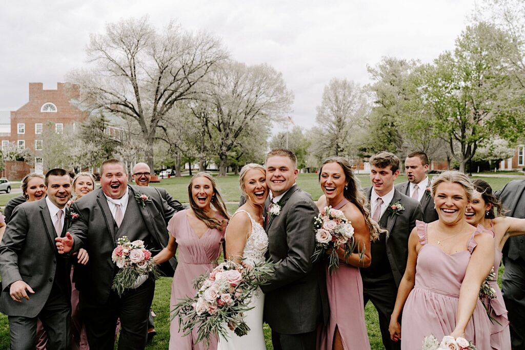 A bride and groom smile at the camera and embrace while their wedding parties smile and swarm them on either side in the middle of a field