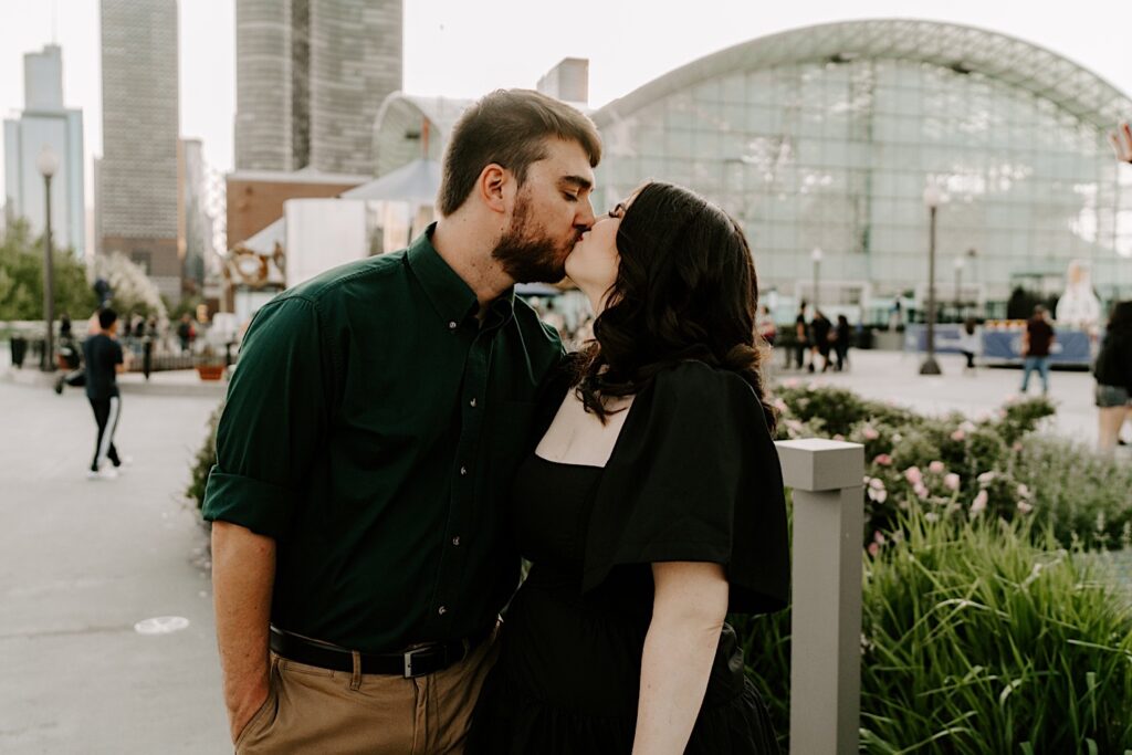A couple kiss one another during their engagement session at Navy Pier in Chicago, behind them is one of the buildings of Navy Pier as well as the Chicago skyline