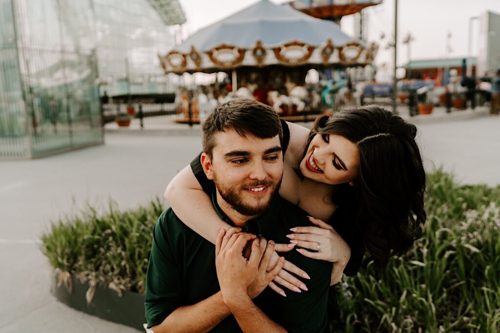 A man smiles as his fiancée smiles and hugs him from behind during their engagement session at Navy Pier in Chicago, behind them is a carousel