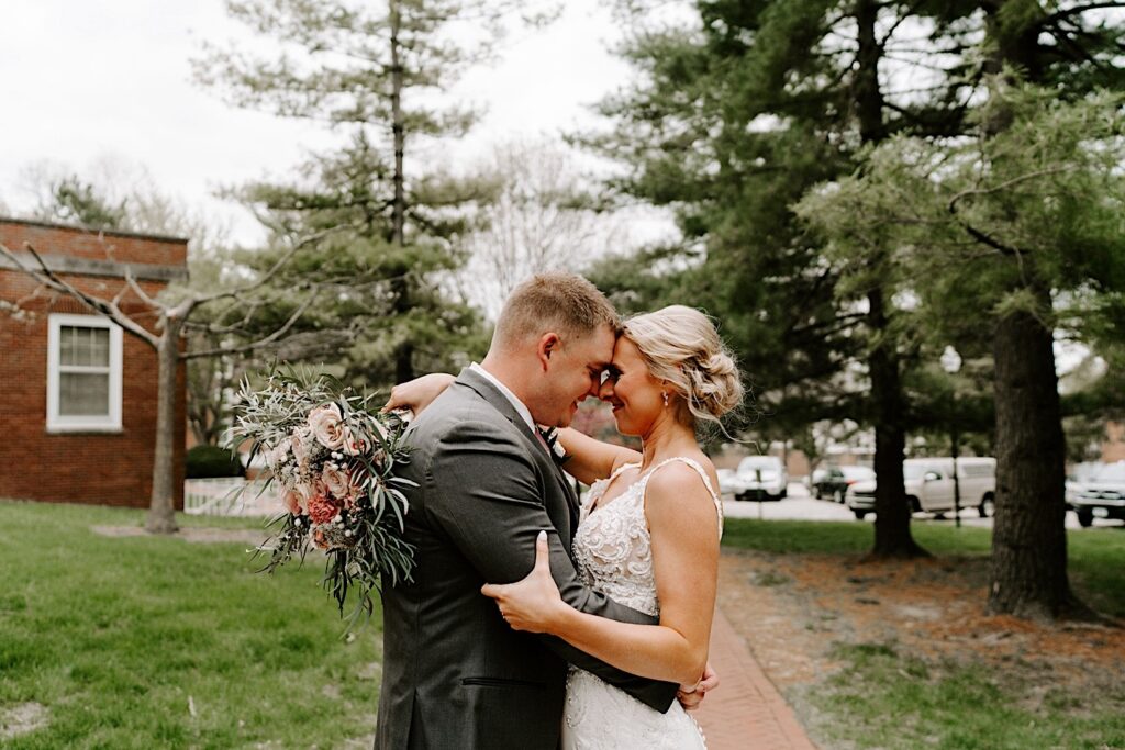 A bride and groom touch foreheads together and smile while standing in a park after their wedding ceremony before heading to their backyard tent reception