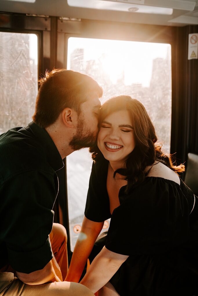 A man kisses his fiancée as she smiles at the camera and the sun sets behind her, they are in a Ferris Wheel cart