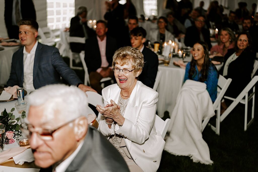A woman laughs and claps while seated at a table during a backyard wedding reception in a tent