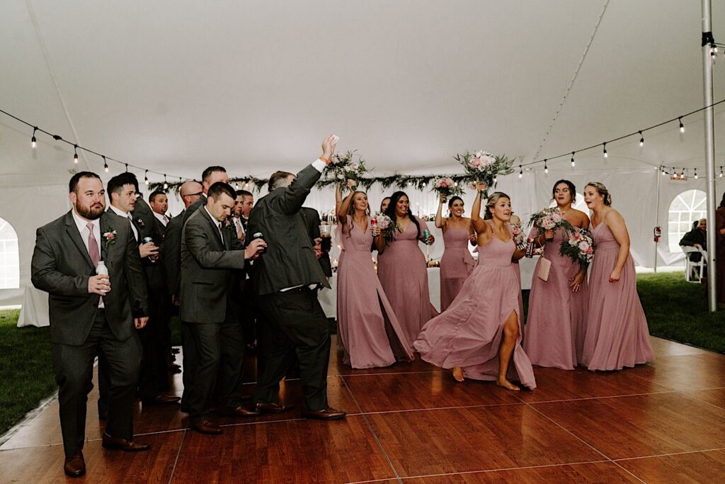 Groomsmen and bridesmaids all stand on a dance floor together and hype up the crowd of a backyard tent wedding reception