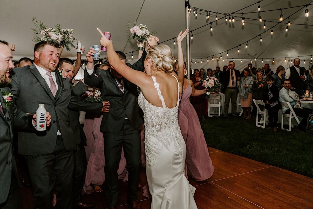 A bride goes in for a hug from her groom who is surrounded on the dance floor by the groomsmen and bridesmaids during their backyard tent wedding reception