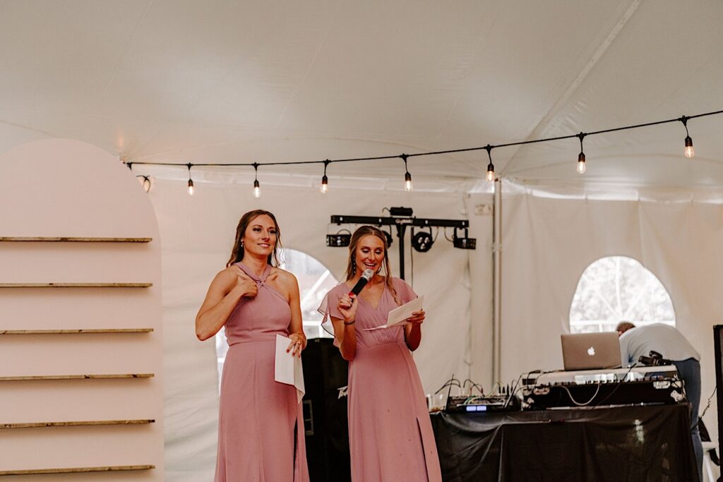 Two bridesmaids stand each holding a piece of paper and one holding a microphone to give speeches during a backyard tent wedding reception