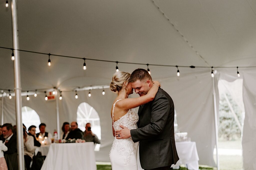 A groom smiles as he and his bride share their first dance together underneath the tent of their backyard wedding reception