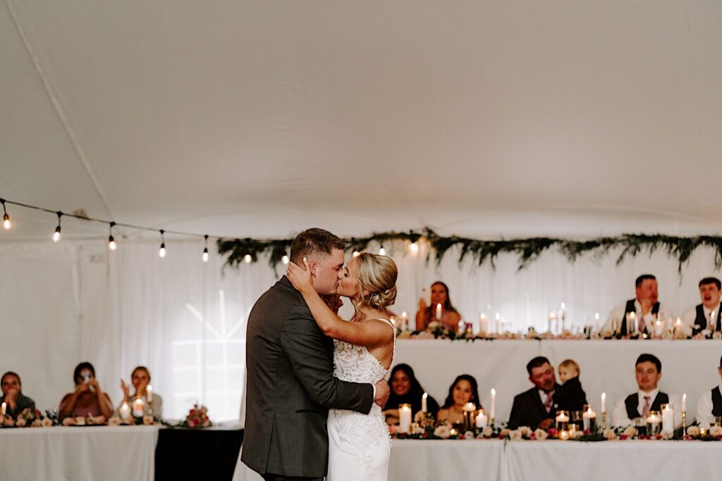 A bride and groom kiss during their first dance during their backyard tent wedding reception as guests behind them cheer and take photos
