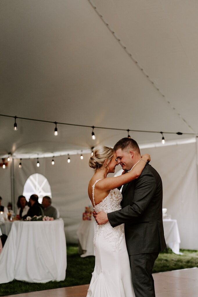 A bride and groom touch foreheads together and smile while doing their first dance underneath a tent