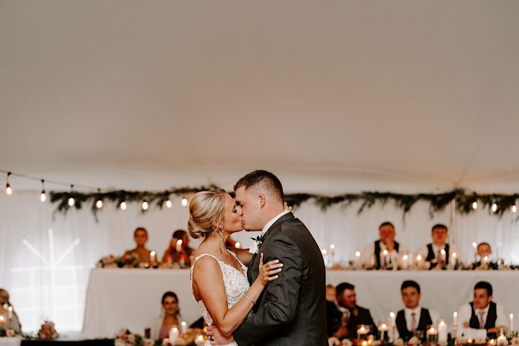 A bride and groom kiss during their first dance during their backyard tent wedding reception as guests behind them watch.