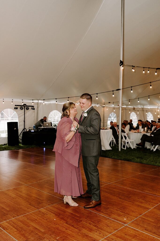 A groom smiles as he dances with his mother on his wedding day underneath a tent