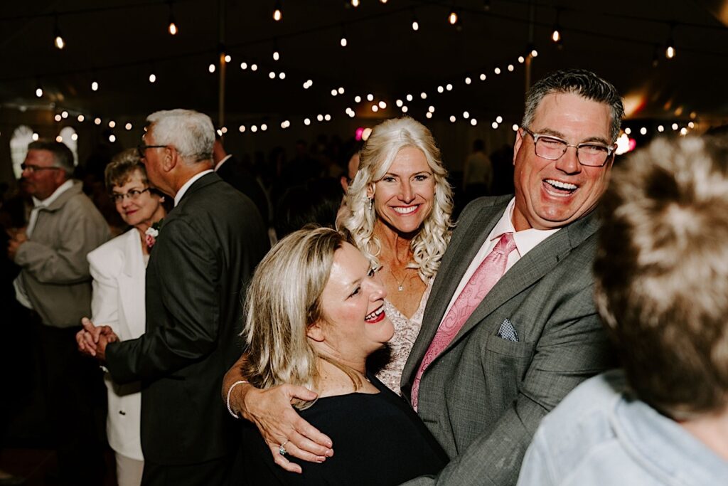 Guests of a backyard tent wedding smile and dance with one another underneath the string lights