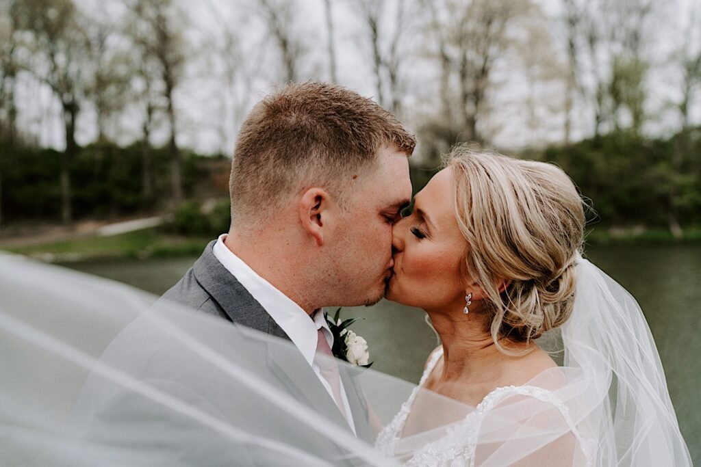 A bride and groom kiss in front of a small lake while the bride's veil flows in the wind in front of them