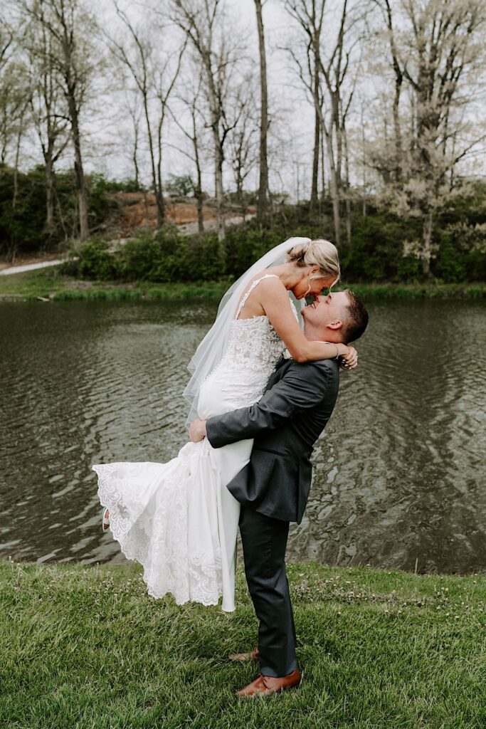 A groom picks up the bride as they smile at one another while standing in front of a small lake
