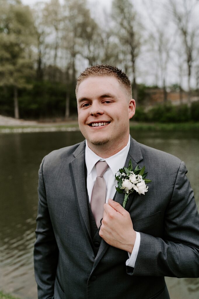 A groom smiles at the camera and adjusts his suit coat in front of a small lake