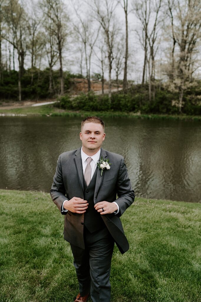 A groom walks towards the camera while adjusting his suit coat in front of a small lake