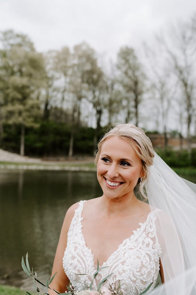 A bride smiles at something off camera while standing in front of a lake