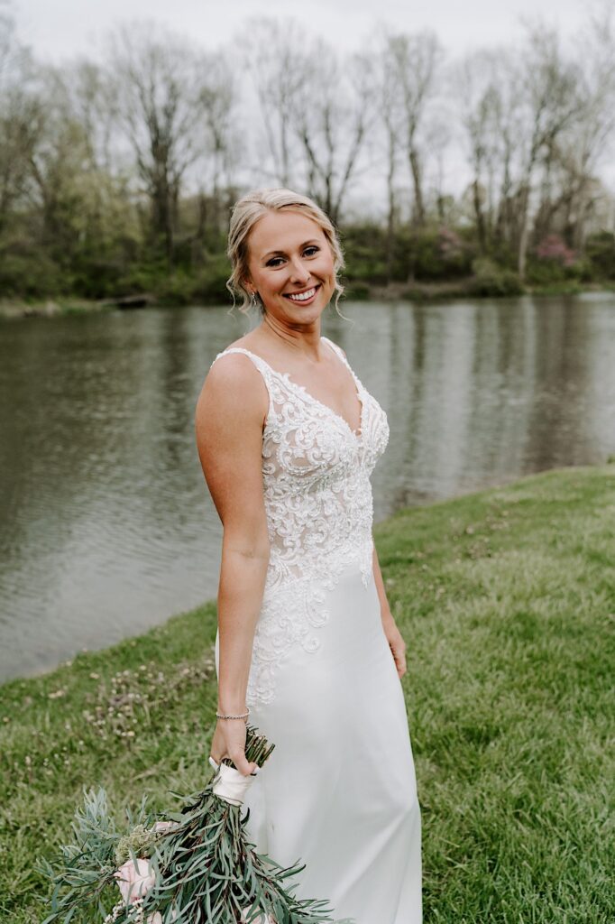 A bride smiles at the camera while holding her bouquet and standing in front of a small lake