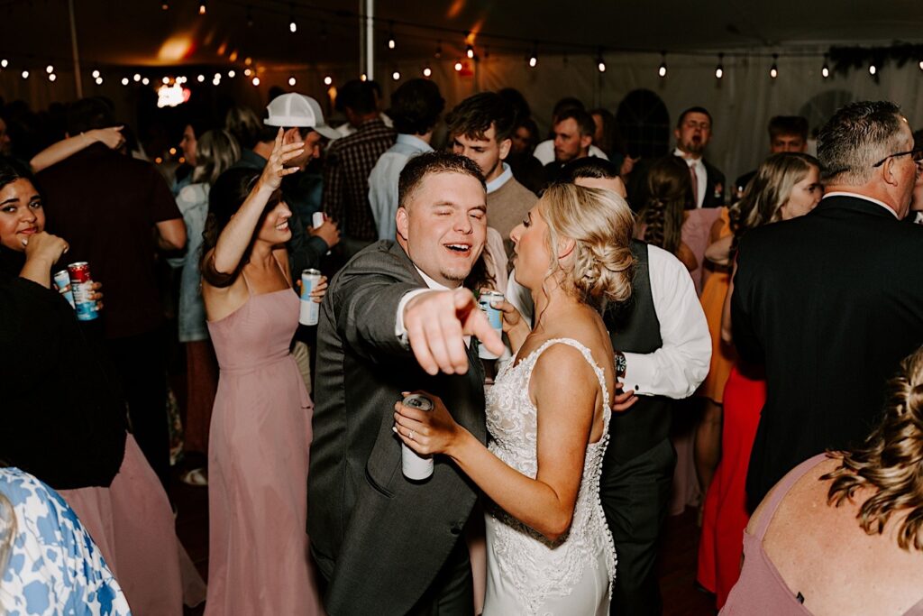 A groom smiles and points at the camera as the bride talks to him and their guests dance around them during their backyard tent wedding reception