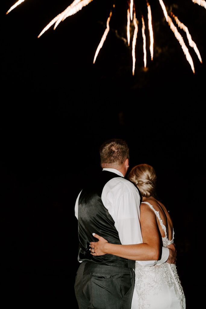 A bride and groom embrace and look up in the sky away from the camera as a firework explodes