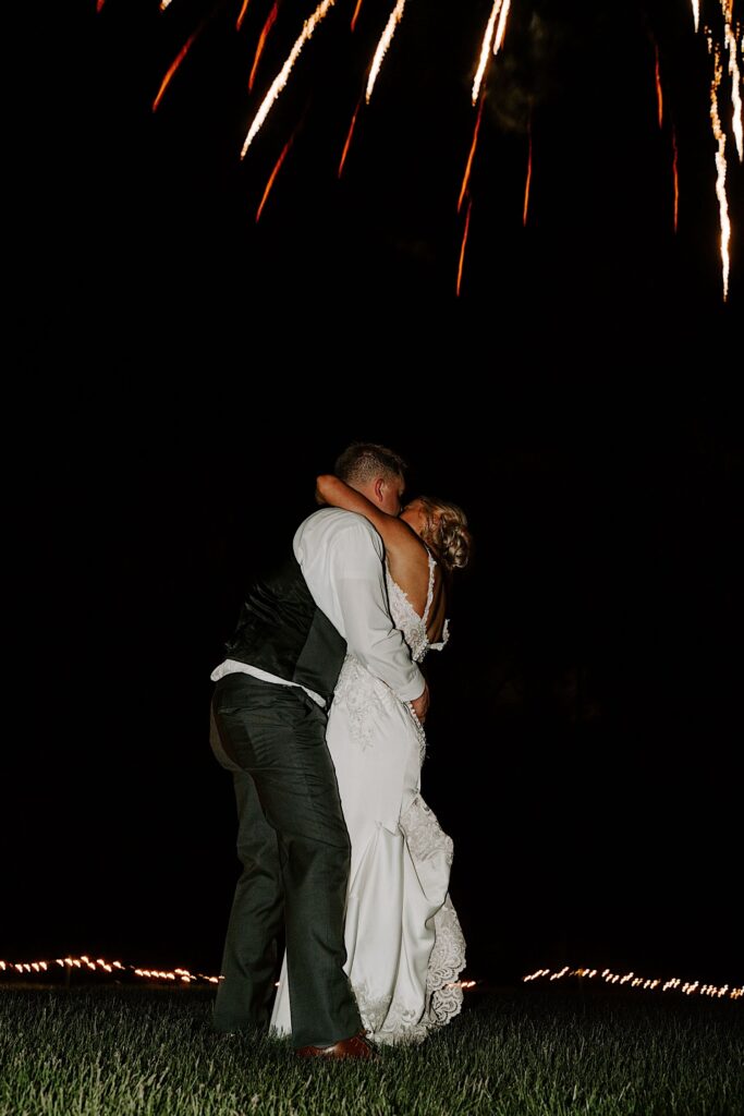 A bride and groom kiss in a field as a firework explodes overhead