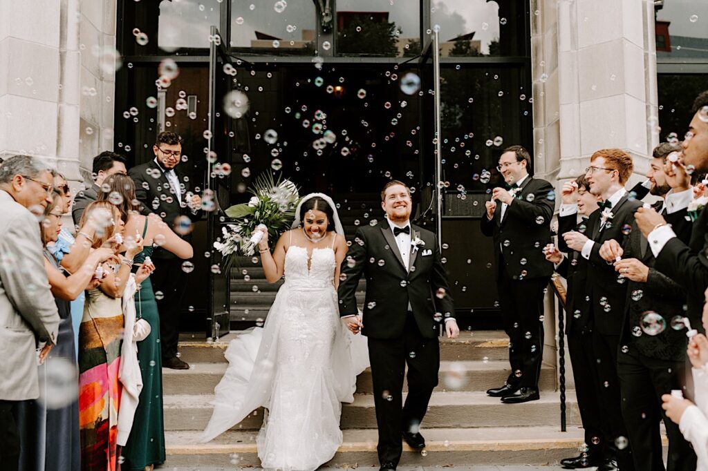 A bride and groom walk down a staircase exiting their wedding ceremony while smiling, on either side of them their guests blow bubbles all around them