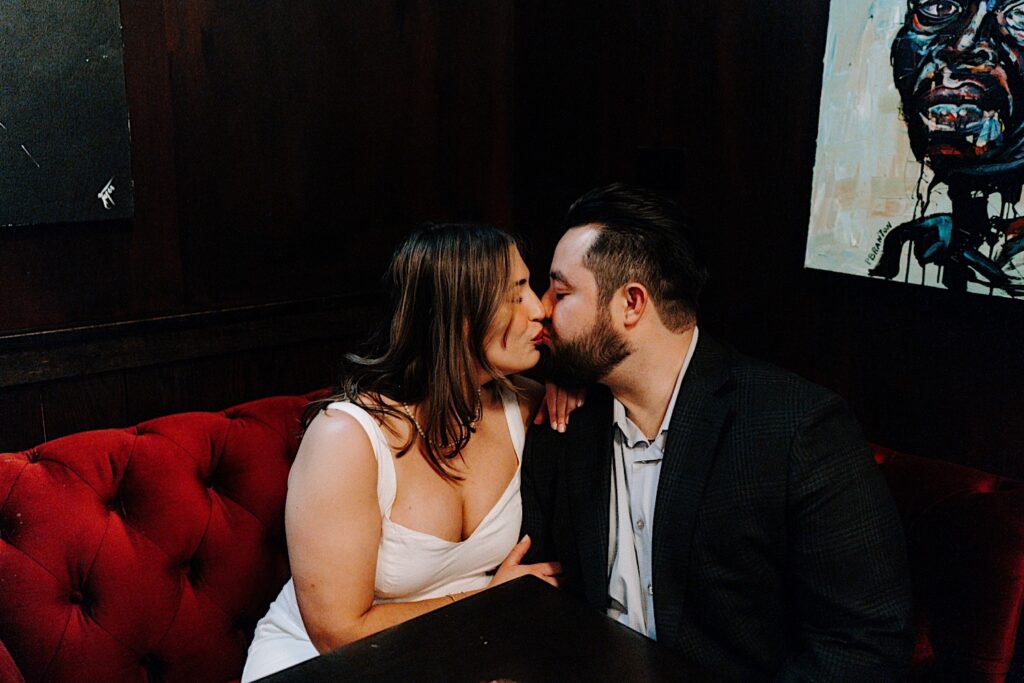 A couple kiss while sitting on a red couch in a dark room during their indoor engagement photos in Chicago