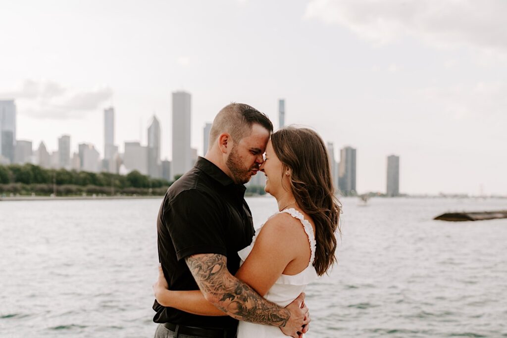 A couple touch their noses together while smiling and laughing in front of Lake Michigan and the Chicago skyline at their engagement session at Museum Campus