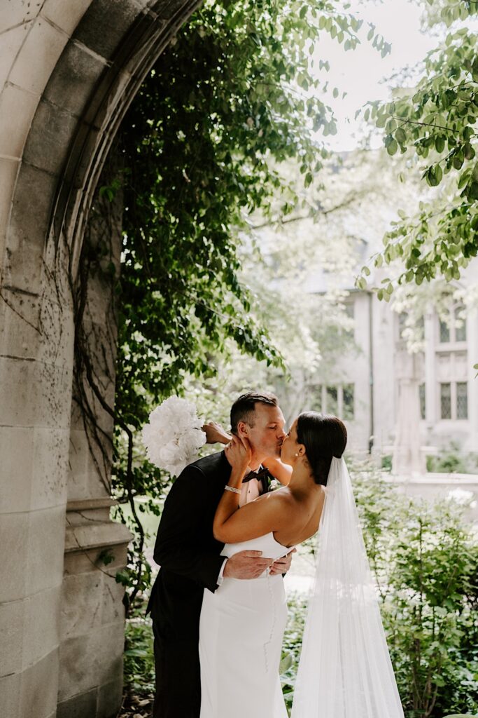 A bride and groom kiss underneath a stone archway with greenery all around them