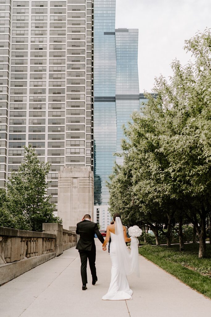 A bride and groom hold hands and walk away from the camera, exiting Millennium Park in Chicago and walking towards the skyscrapers