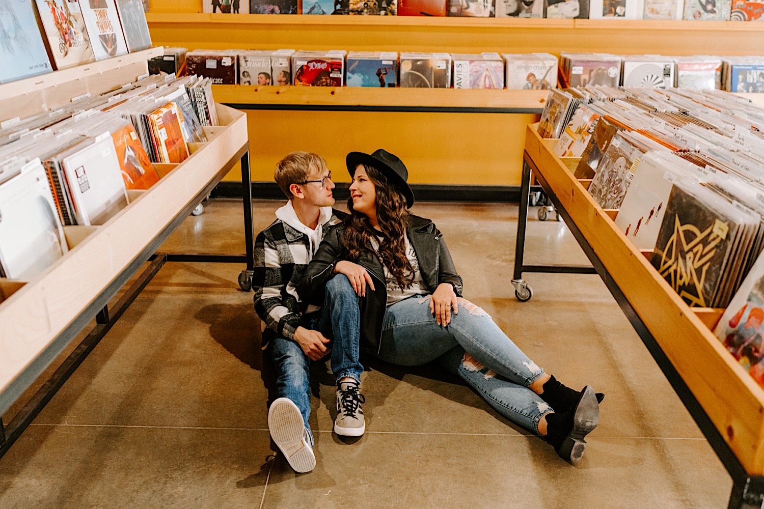 A couple sit on the ground indoors at a record store in Chicago and look at one another while taking engagement photos