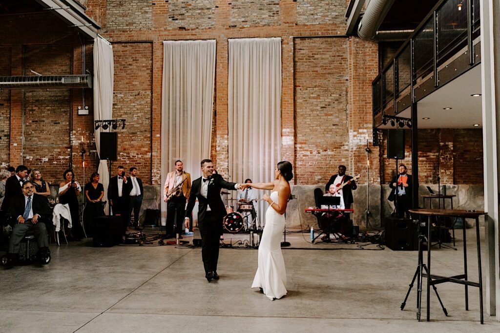 A bride and groom dance in a large brick building with a live band playing behind them and their guests to the left cheering for and watching them