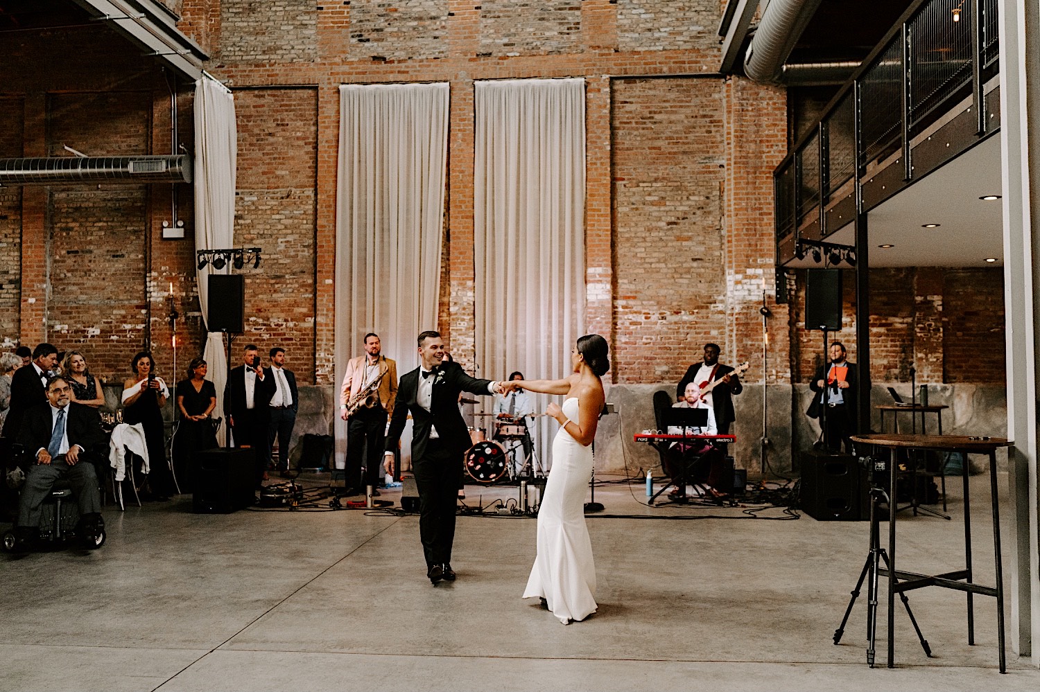 A bride and groom dance in a large brick building with a live band playing behind them and their guests to the left cheering for and watching them
