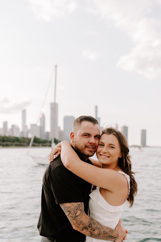 A couple embrace and touch their cheeks together while smiling at the camera in front of Lake Michigan and the Chicago skyline