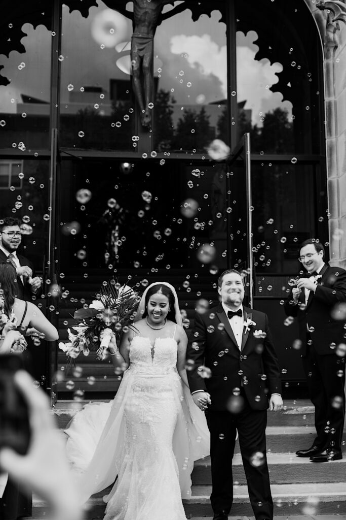 Black and white photo of a bride and groom smiling walking out of a building as wedding guests blow bubbles on either side of them