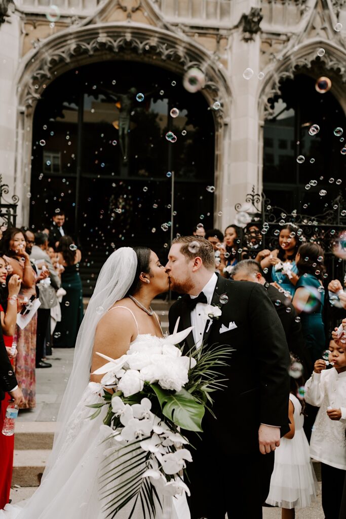 A bride and groom kiss while walking down stairs exiting the building of their wedding reception, on either side of them guests are blowing bubbles all around them