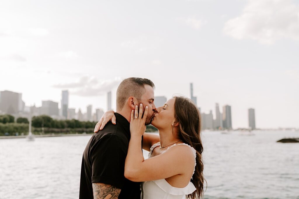 During their engagement session at Museum Campus, a couple kiss each other in front of Lake Michigan and the Chicago skyline while the woman has her hand on the man's face showing off the engagement ring