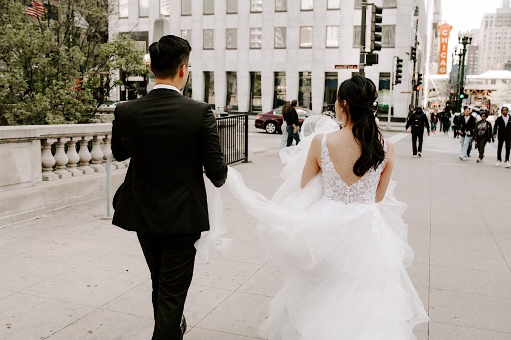 A bride and groom walk with one another and each hold part of the bride's dress as they walk away from the camera along a sidewalk in Chicago