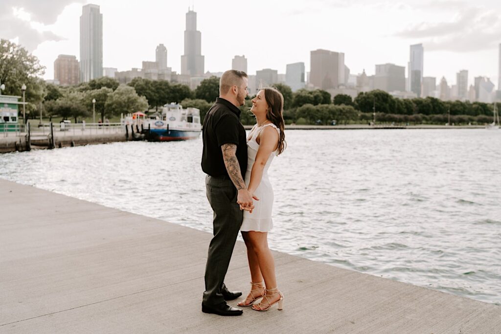 During their engagement session at Museum Campus, a couple stand together and hold hands while smiling at each other in front of Lake Michigan and the Chicago skyline