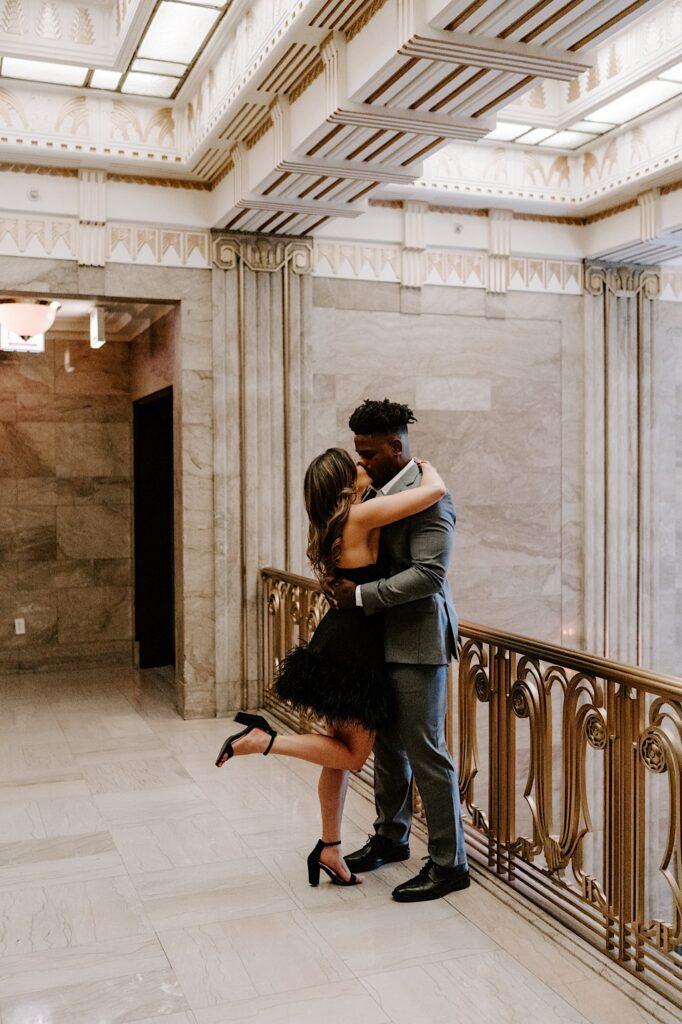 In a building with marble walls and floors a man and woman kiss while hugging next to a golden railing on the balcony